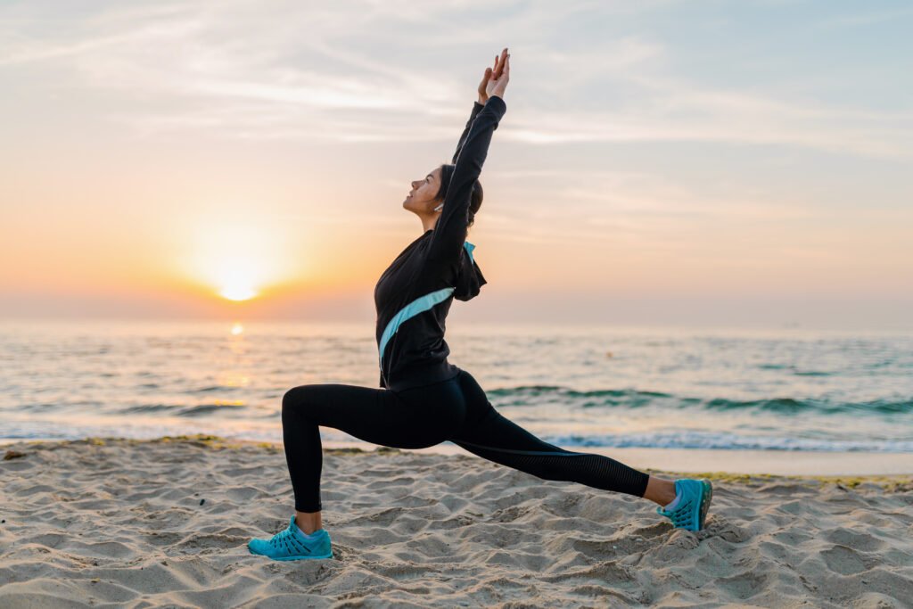 young attractive slim woman doing sport exercises on morning sunrise beach in sports wear, healthy lifestyle, listening to music on earphones, making yoga